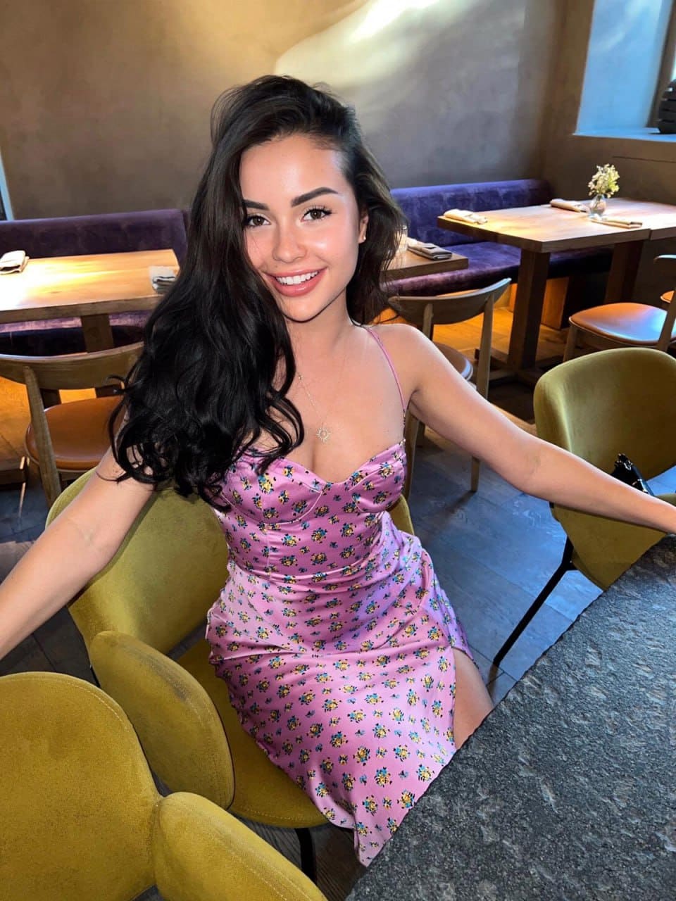 A woman in a pink floral mini dress sits smiling in a mustard-yellow chair at a restaurant table.