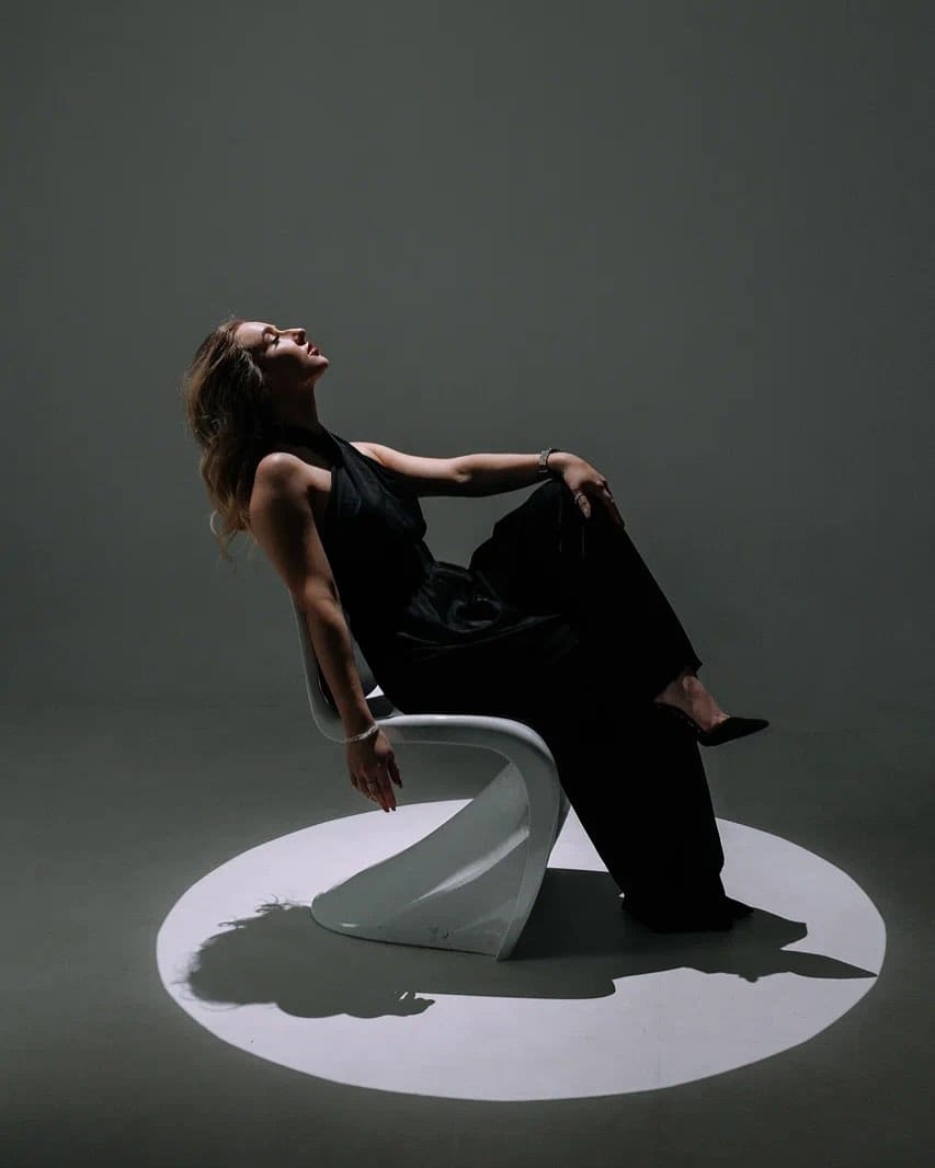 A woman sits on a white sculptural chair in a black top and trousers, head tilted back, legs crossed, in a dim studio with a floor spotlight.