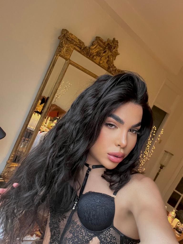 A woman with long dark hair poses in a black lace bodysuit with a halter neck, standing in front of a large ornate gold-framed mirror indoors.