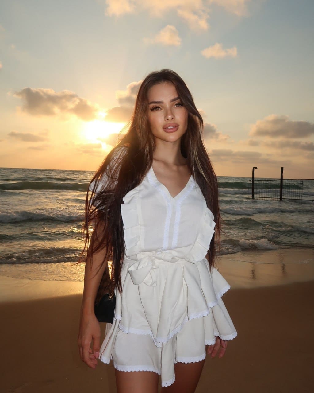 A woman stands on a sandy beach at sunset in a white ruffled top and shorts with a tied waist, facing the camera with ocean waves behind her.