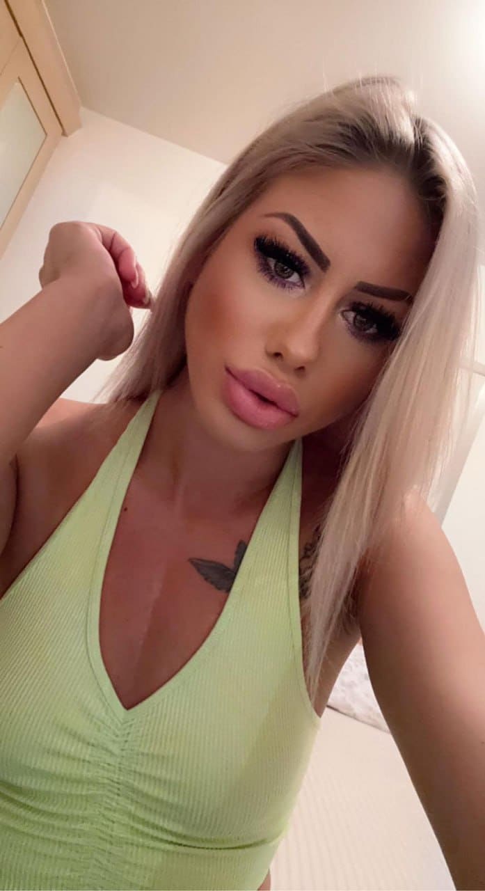 Close-up selfie of a blonde woman in a light green halter top, hand raised near her shoulder, revealing a chest tattoo.