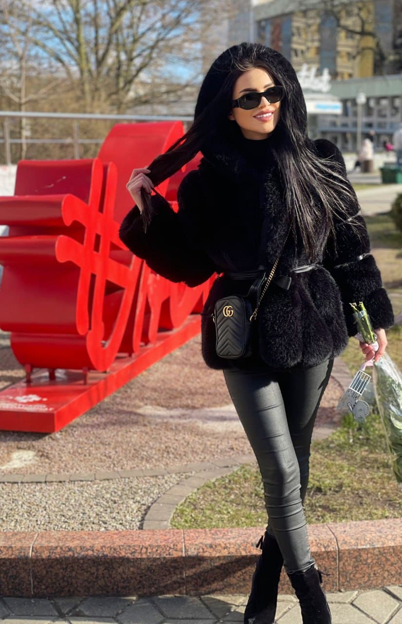 Woman in a black fur coat, turtleneck, and boots stands outdoors holding a bouquet and bag, in front of a large red sculpture.