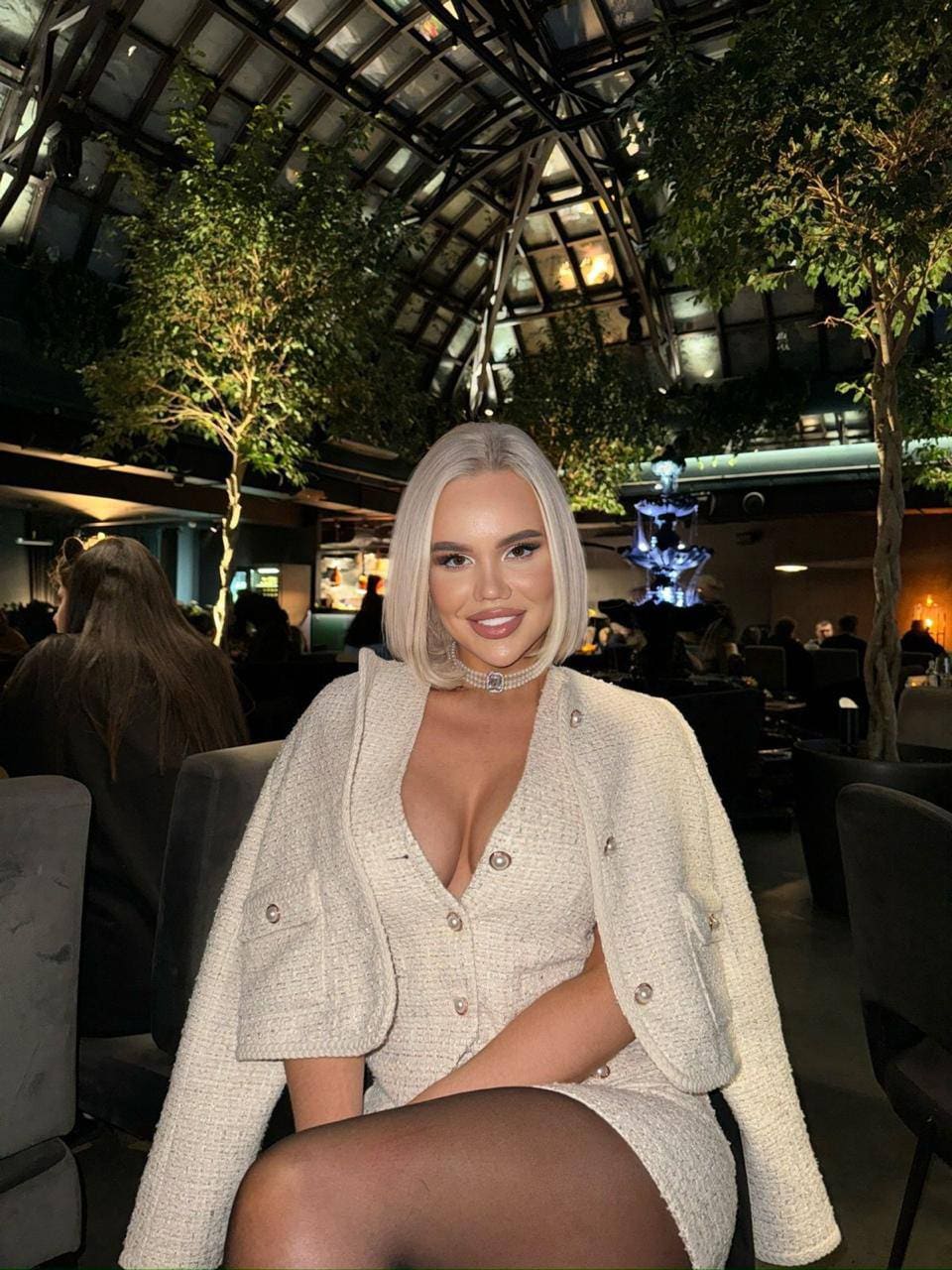A blonde woman in a cream tweed outfit and pearl choker sits in a lounge with a glass-domed ceiling and indoor trees.