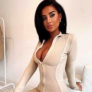A woman sits indoors wearing a ribbed cream long-sleeve top with a deep V-neck and zipper, against a light wall with a white chair.