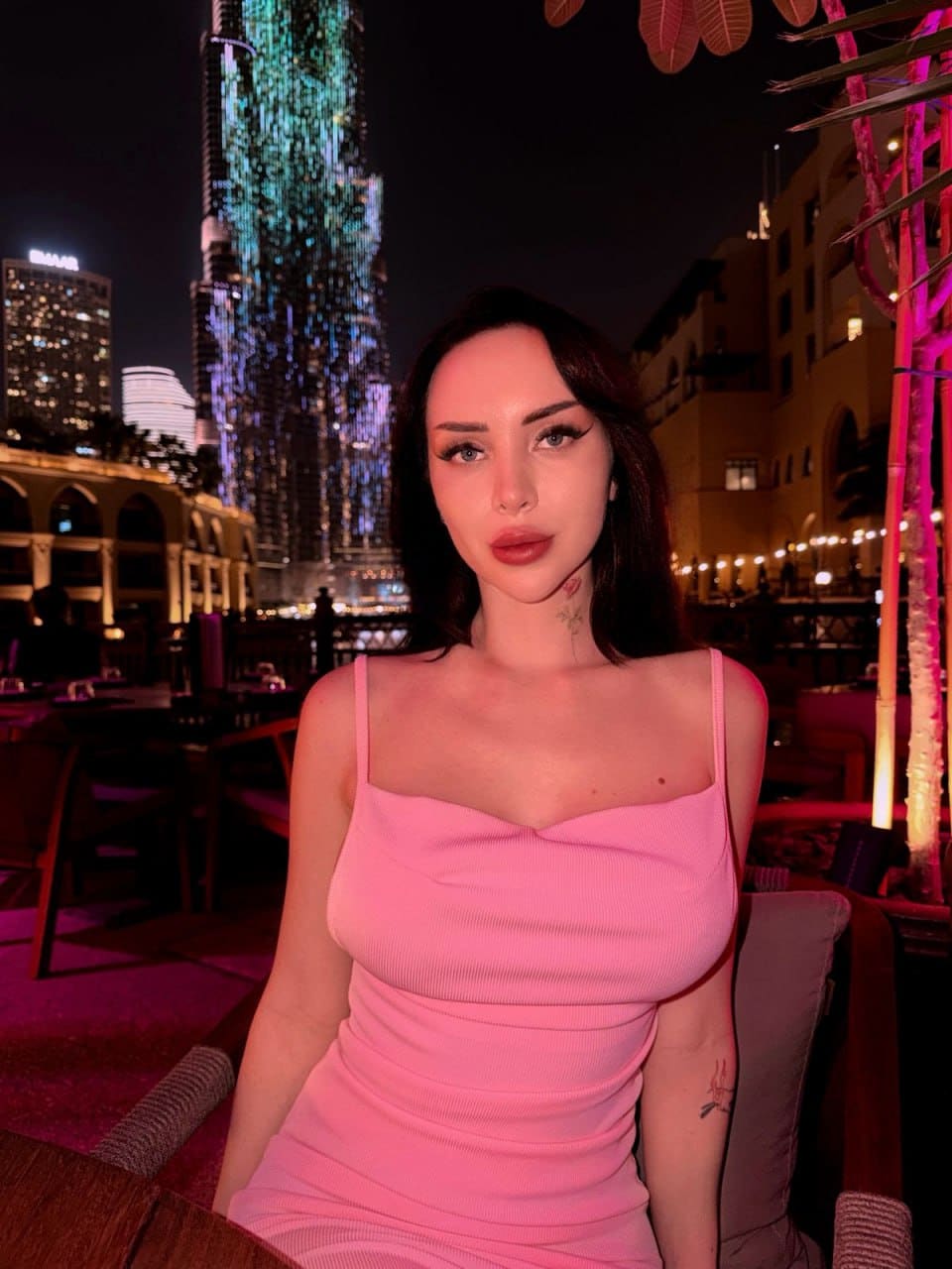 A woman in a pink ribbed dress sits at night, with a lit skyscraper and palm trees visible in the background.