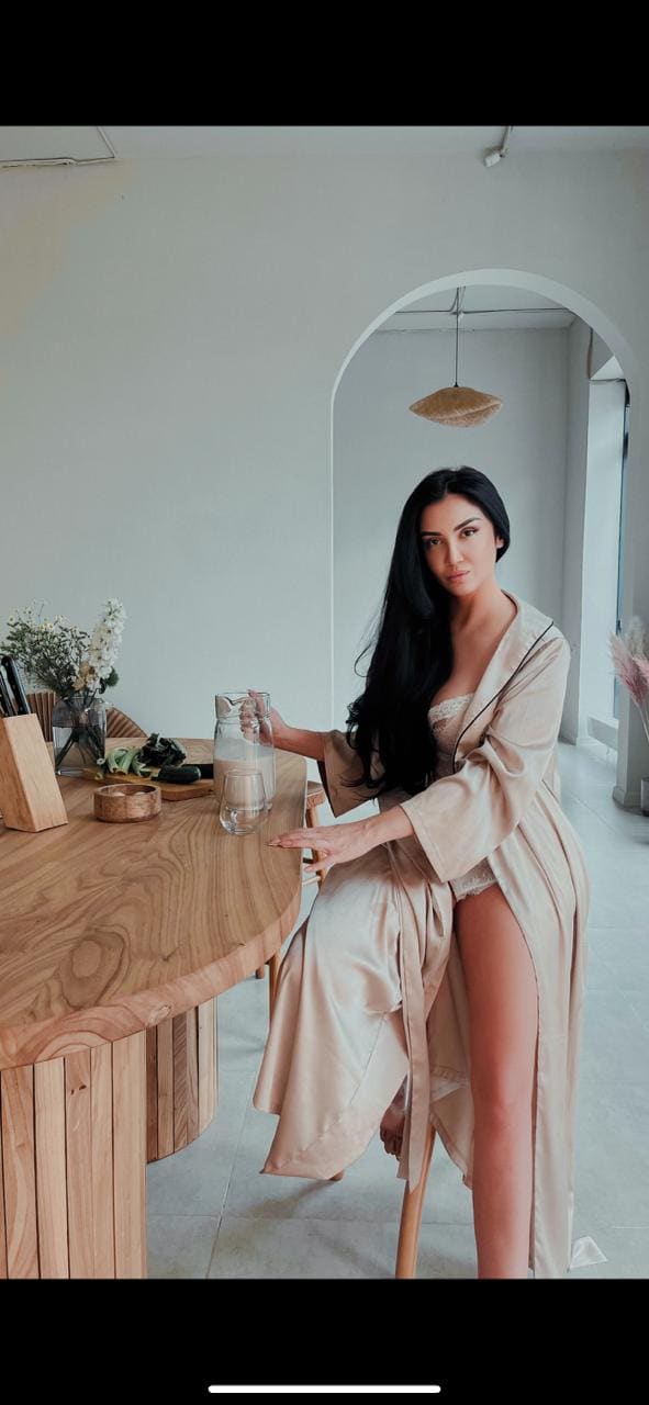 A woman sits at a wooden table in a beige robe over lace underwear, one hand resting on a glass of water.
