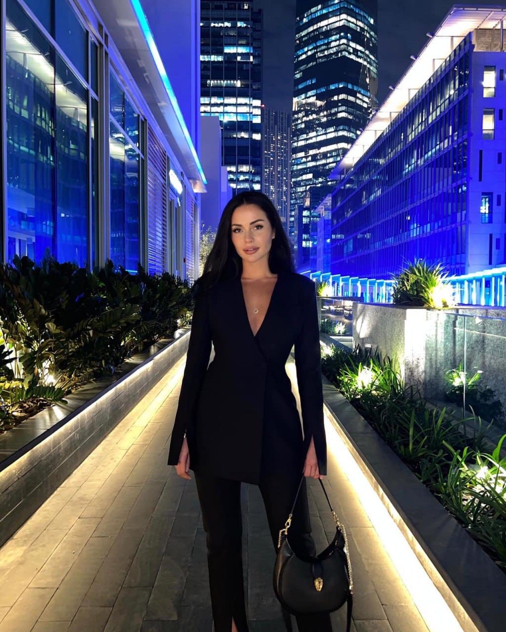 A woman in a black blazer dress and trousers holds a handbag on a lit walkway at night, with glass buildings in the background.