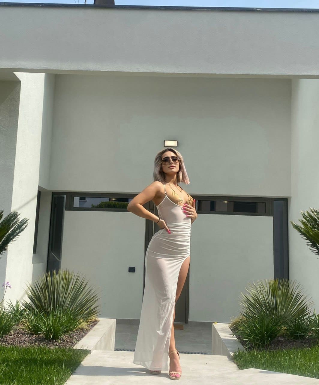 A woman in sunglasses and a long white slit dress stands on a walkway in front of a modern white building.