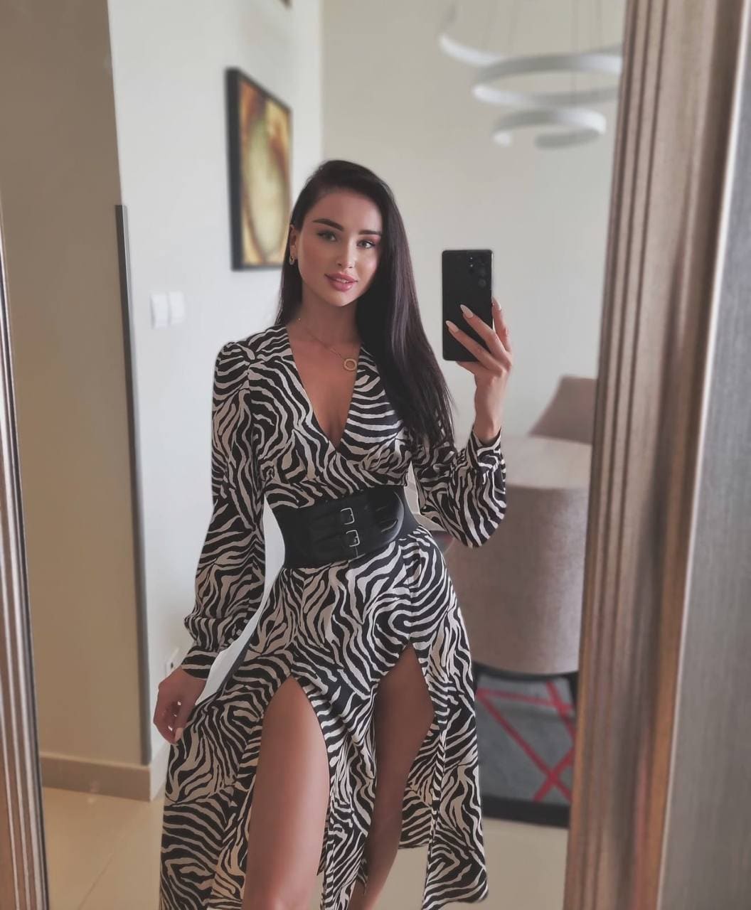 A woman takes a mirror selfie in a zebra-print wrap dress with a thigh slit and wide belt in a modern interior with a chandelier.