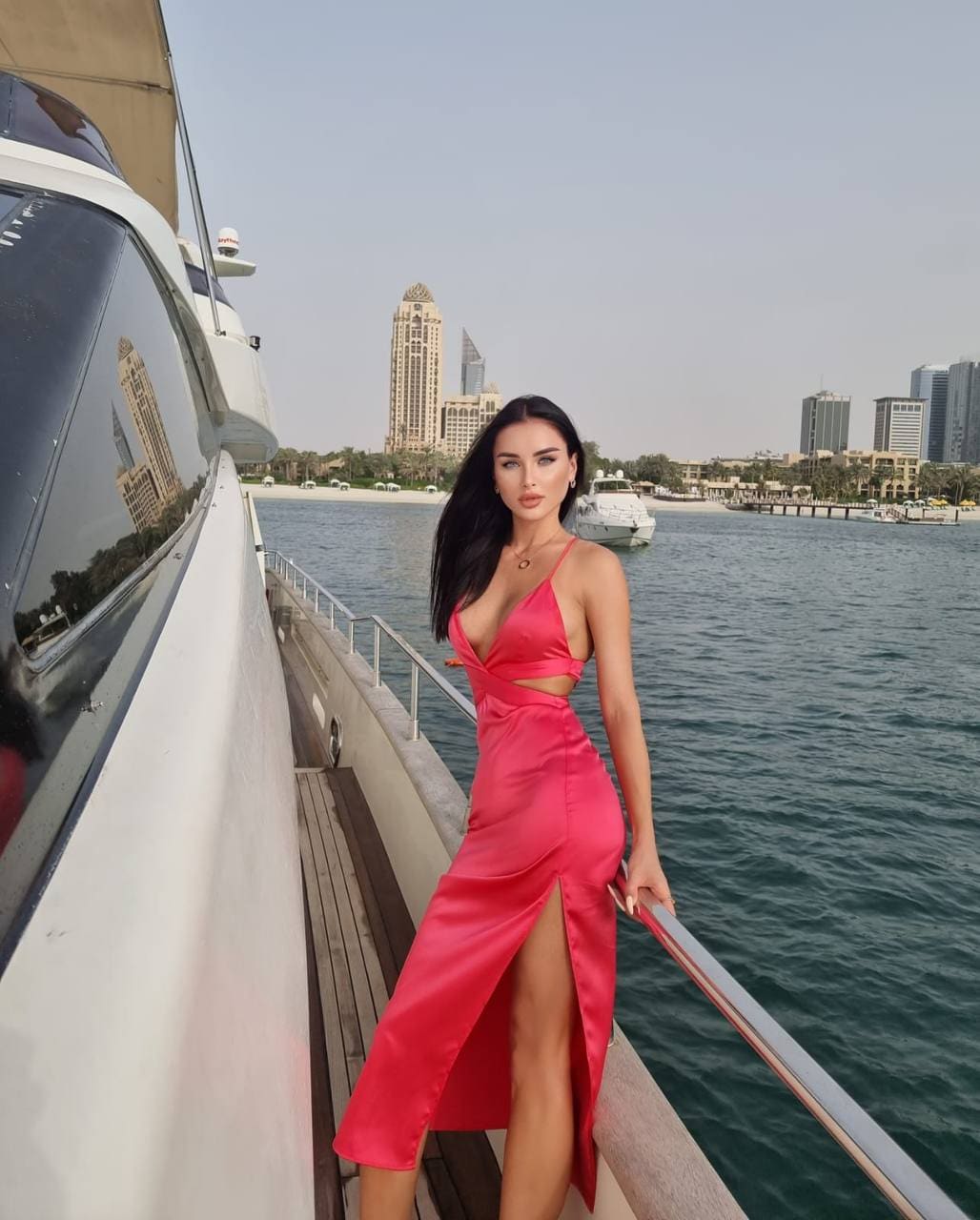 A woman in a bright pink satin dress with a thigh slit stands on a white boat deck, holding the railing against a city skyline.