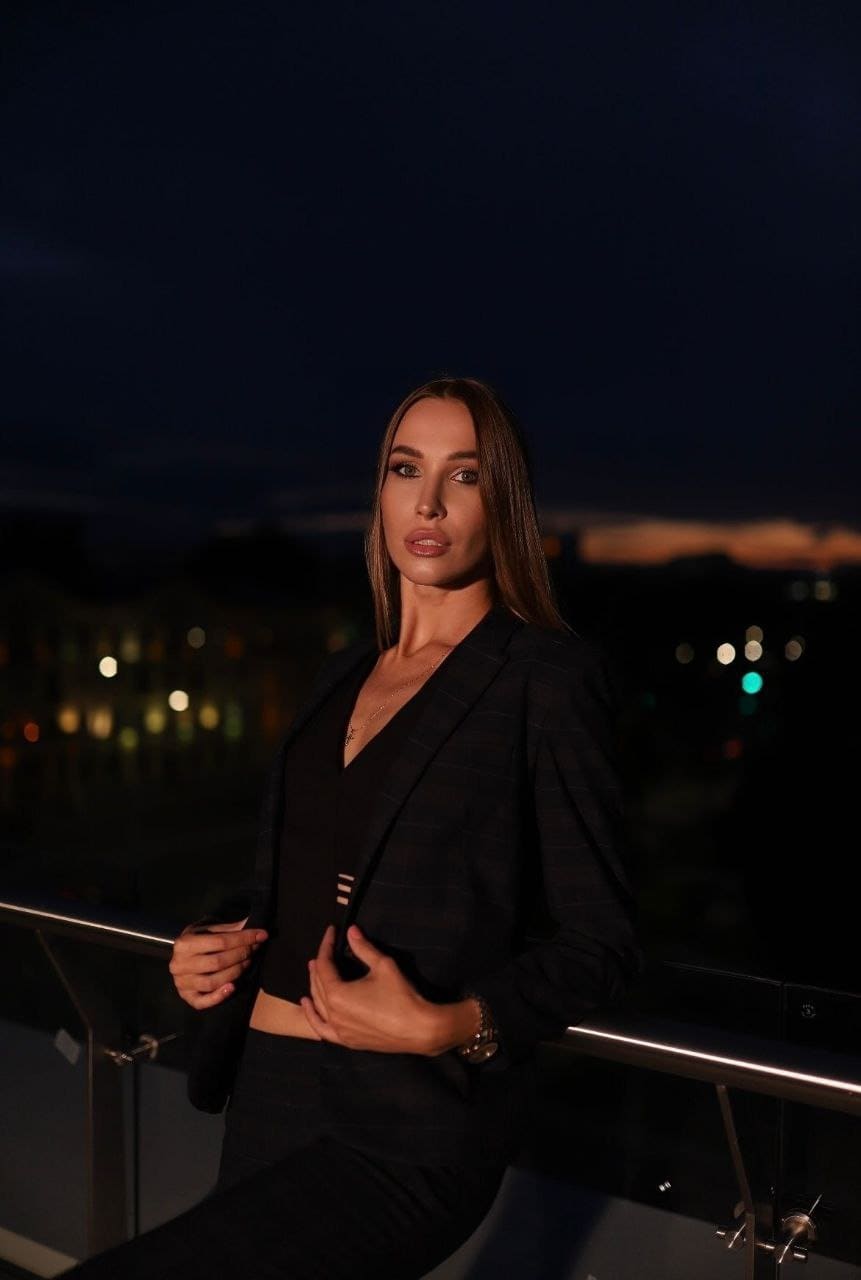 A woman in a dark plaid blazer and black crop top stands on a rooftop terrace at night, one hand on the railing, against blurred city lights.