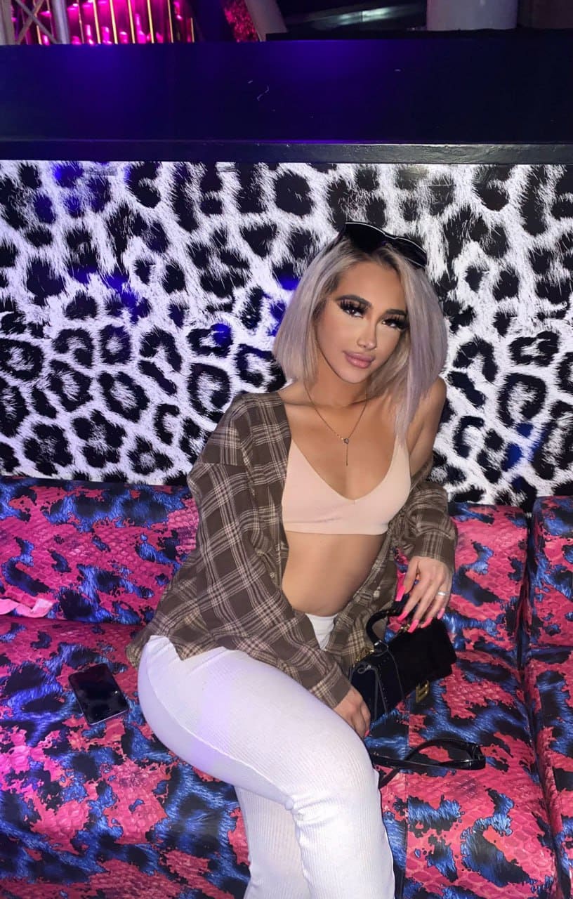 A woman sits on a patterned couch wearing a beige bralette, open plaid shirt, and white pants with a black bag.