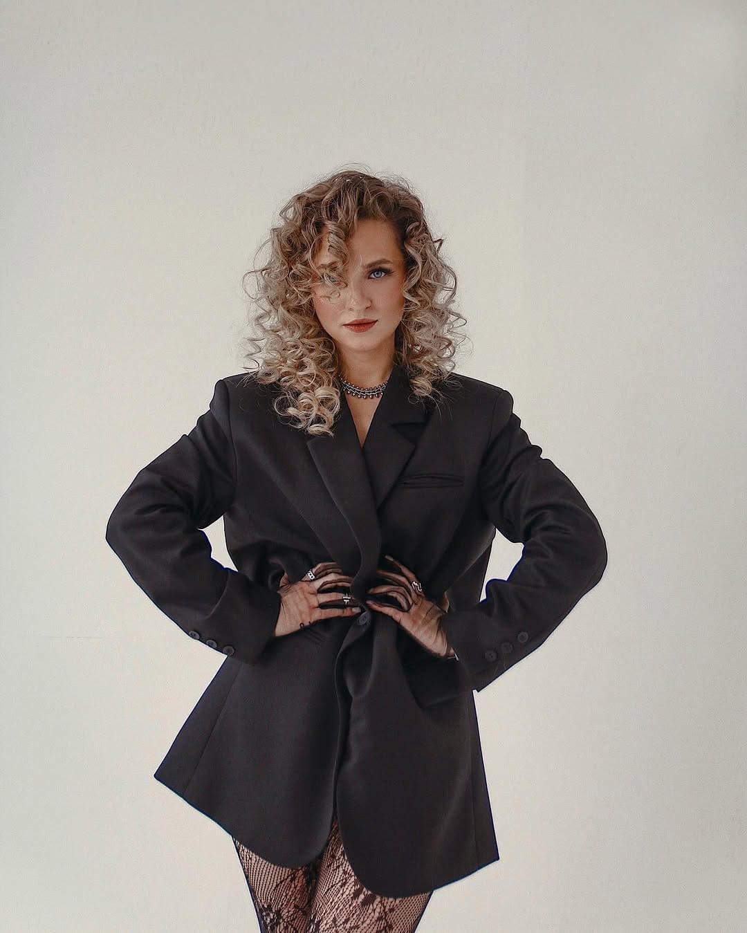 A woman stands with hands on hips wearing a black blazer, dark top, and fishnet stockings against a plain light background.