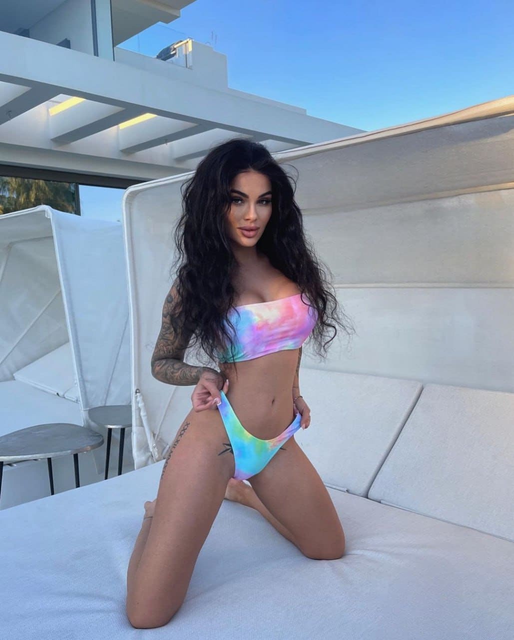 A woman with wavy hair and tattoos kneels on an outdoor lounge in a pastel tie-dye bikini, blue sky behind.