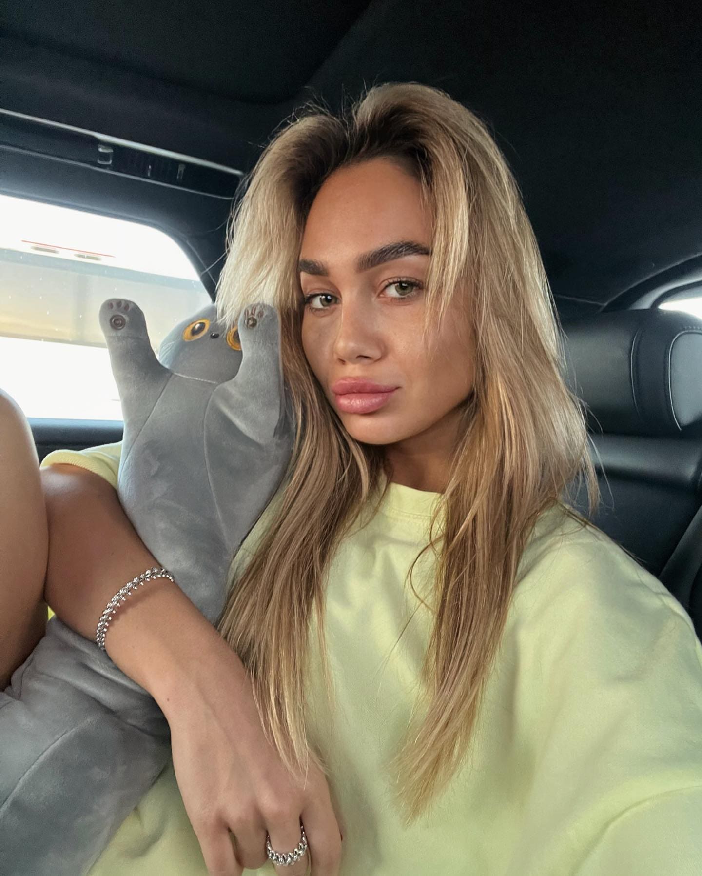 Alesya sits in a car back seat wearing a pale yellow t-shirt, holding a grey plush toy against her shoulder.