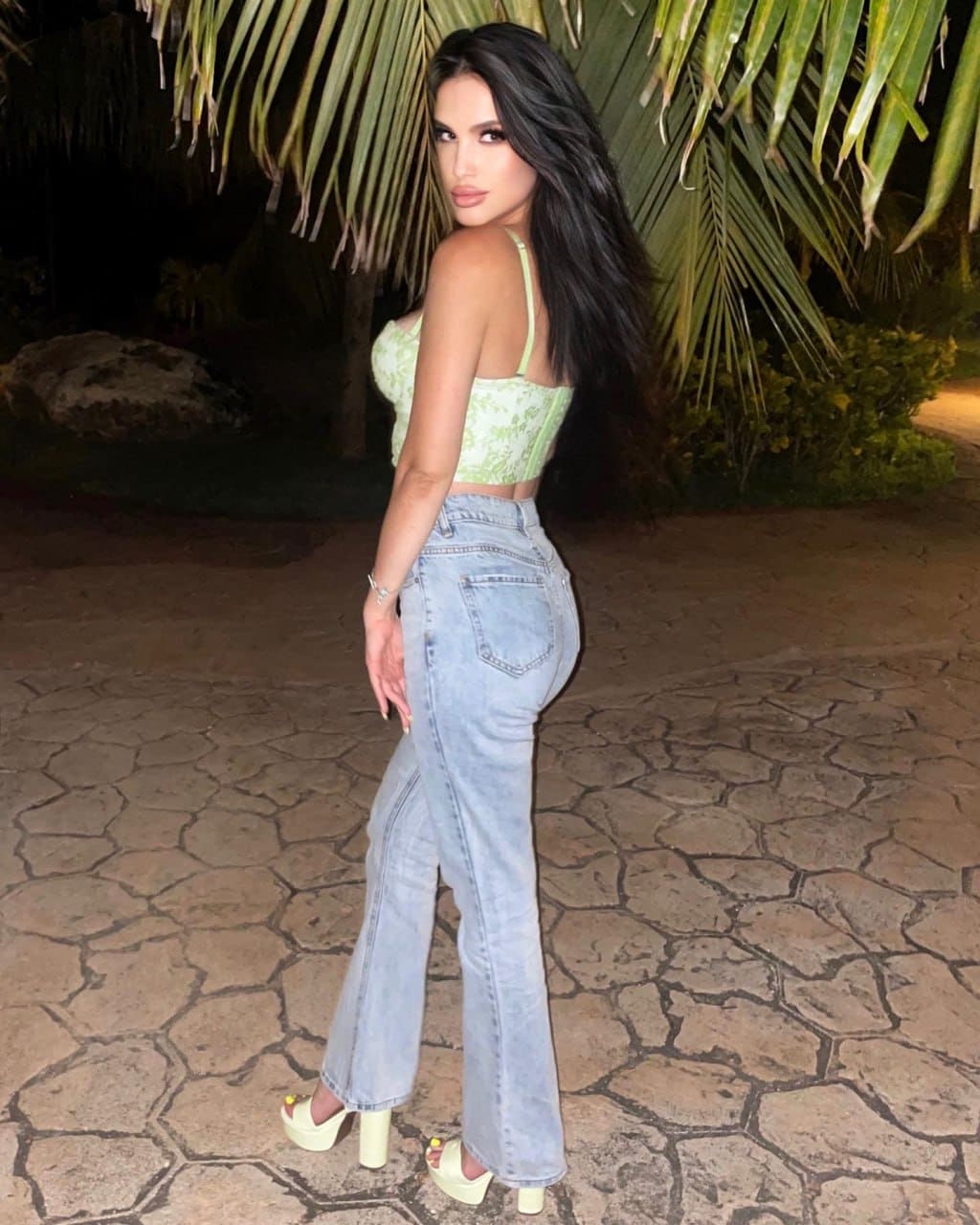 Woman in profile on a stone patio at night, wearing a light green lace crop top, high-waisted jeans, and platform sandals near palm fronds.