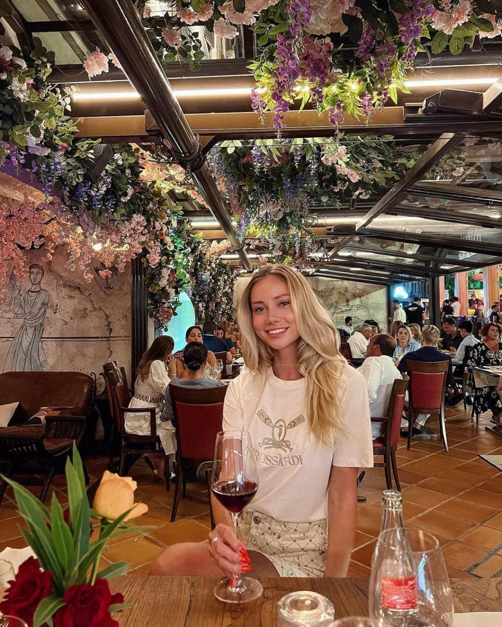 A woman sits at a restaurant table in a white t-shirt and shorts, holding a glass of red wine.