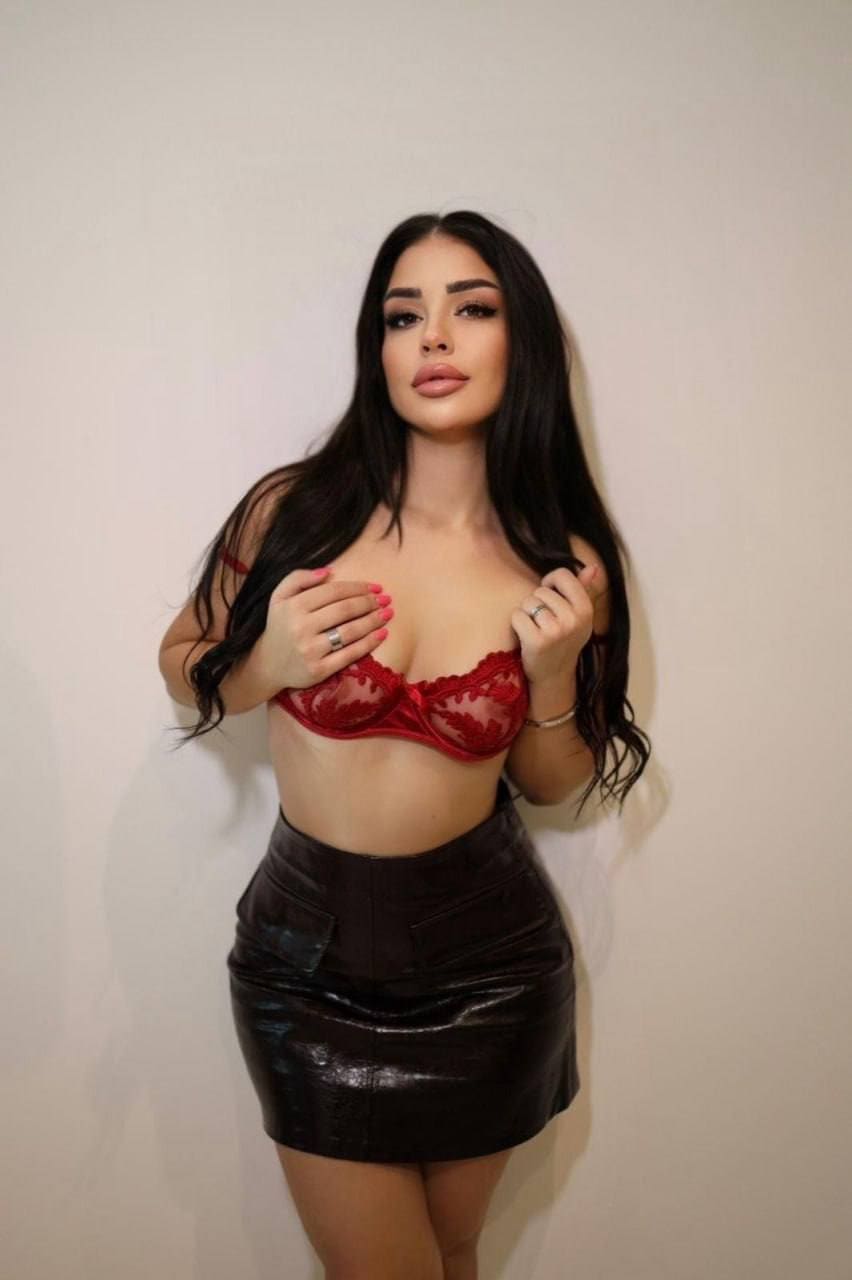 Lillian stands against a light wall in a red lace bra and black leather skirt, hands raised near her chest.