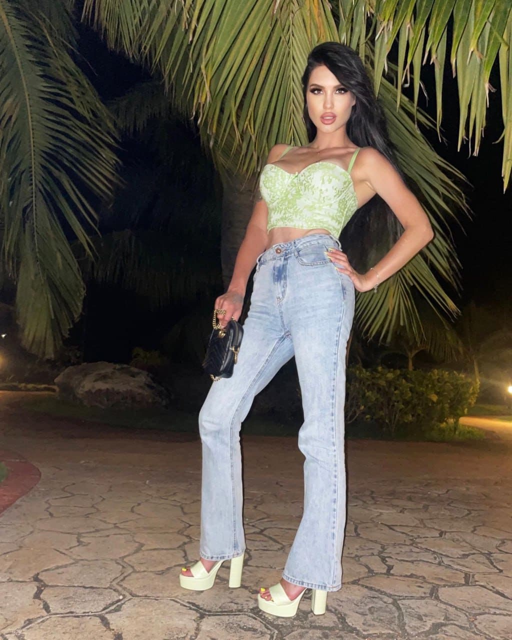 Woman stands on a stone path at night in a light green crop top, flared jeans, and cream sandals, holding a small black handbag.