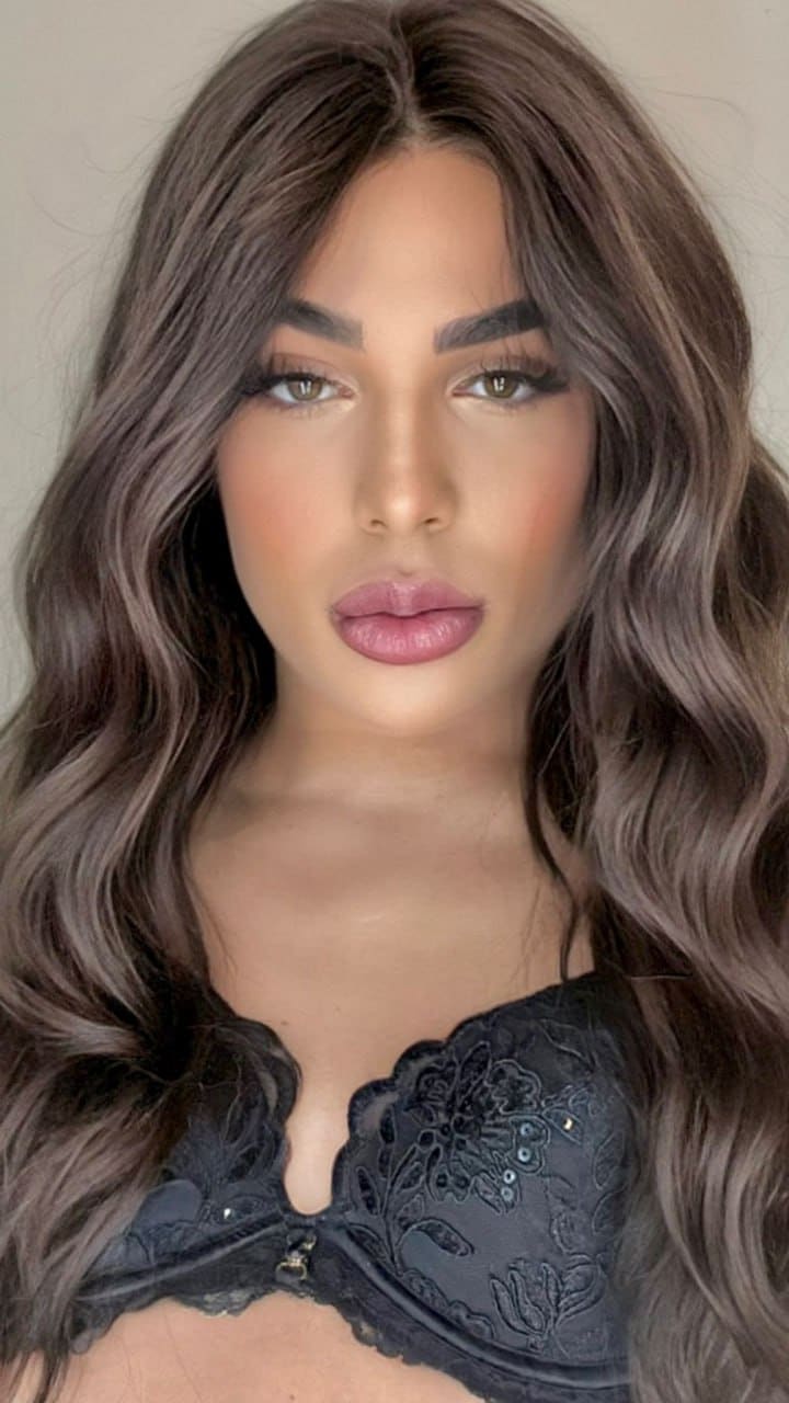 A woman with long brown hair and full makeup faces the camera wearing a black lace bralette, set against a plain light background.