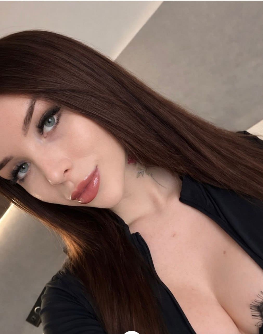 A close-up selfie shows a woman with blue eyes in a black deep-neck top, with a small neck tattoo against a light wall.