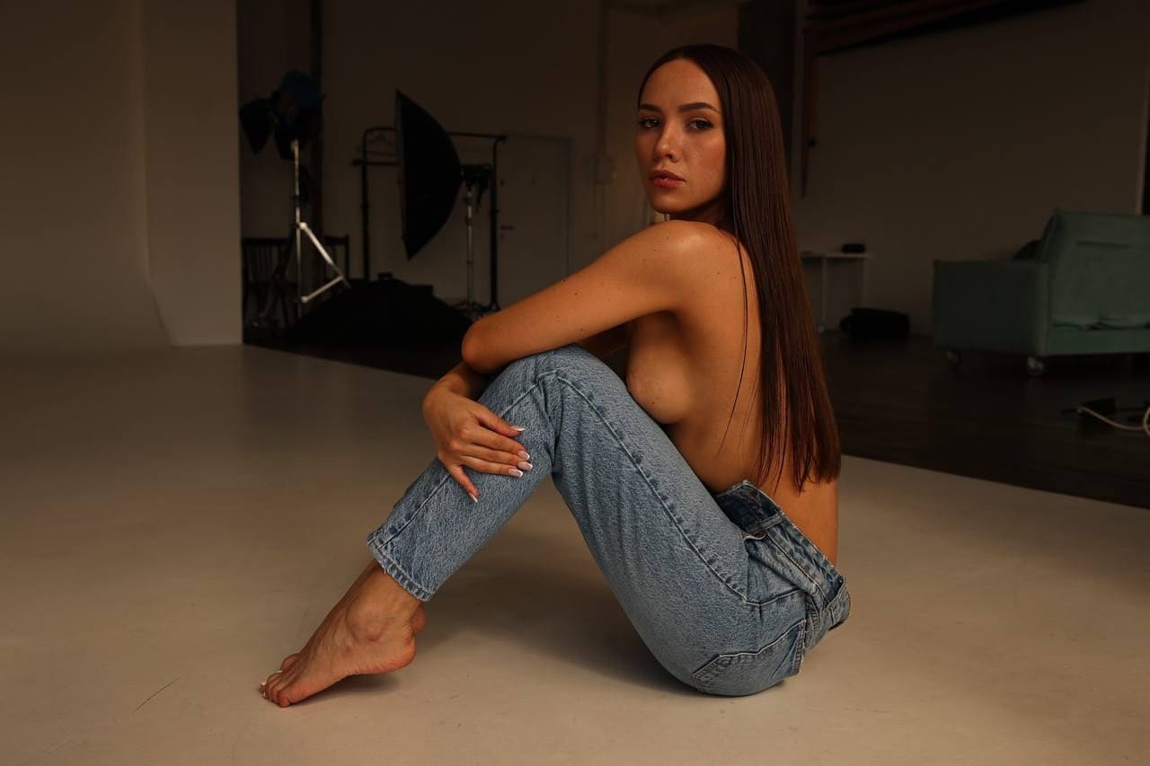 Sitting barefoot on a studio floor in light blue jeans, arms wrapped around knees, with photography equipment in the background.