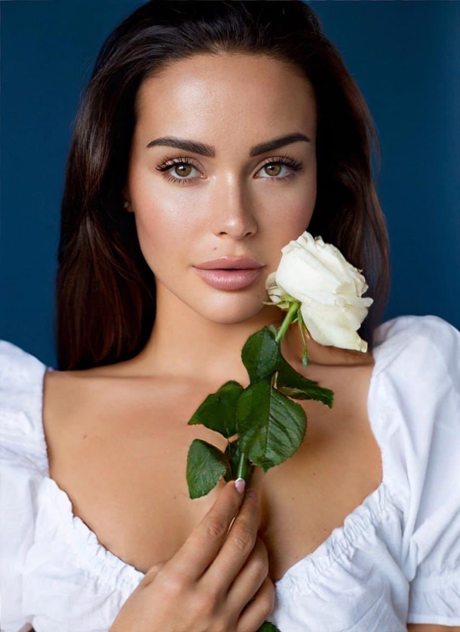 Evangelina holds a white rose near her face, wearing a white off-the-shoulder top with a low neckline against a solid blue background.