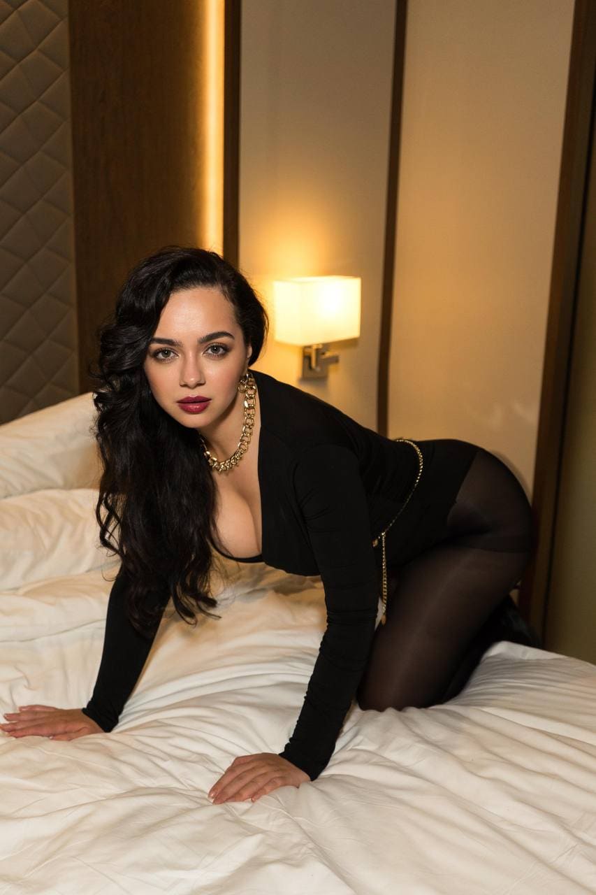 A woman kneels on a bed in a black long-sleeved top and sheer tights, with a gold necklace and wall lamp.