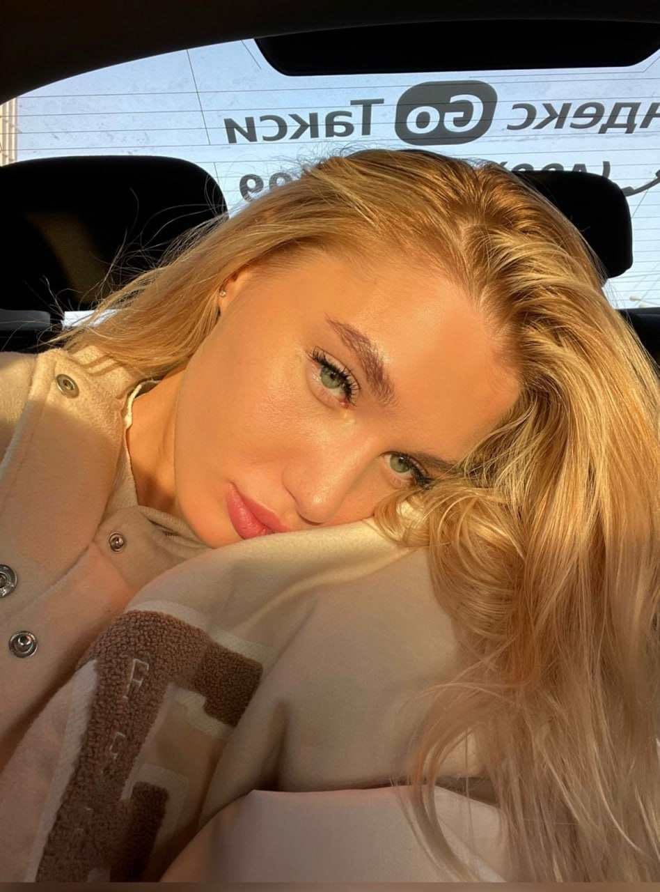 Stella rests her head on her arm while seated in a car, wearing a light buttoned jacket over a patterned top.