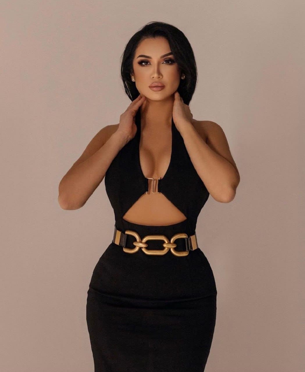 Kamila stands against a beige background in a black halter dress with a gold-buckled belt, hands raised to her neck.