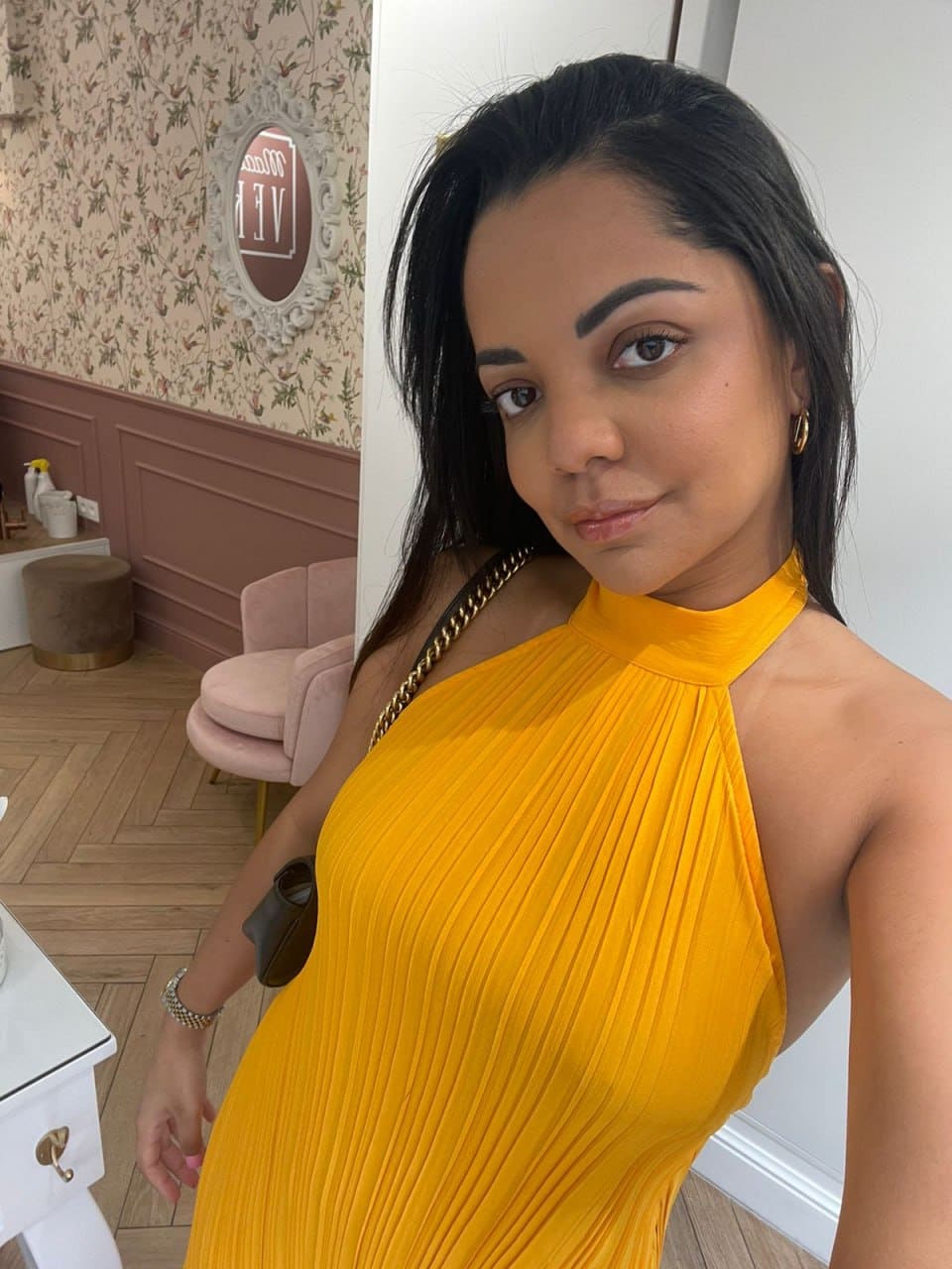 A woman in a yellow pleated halter dress and gold chain bag stands in a room with floral wallpaper and a pink chair.