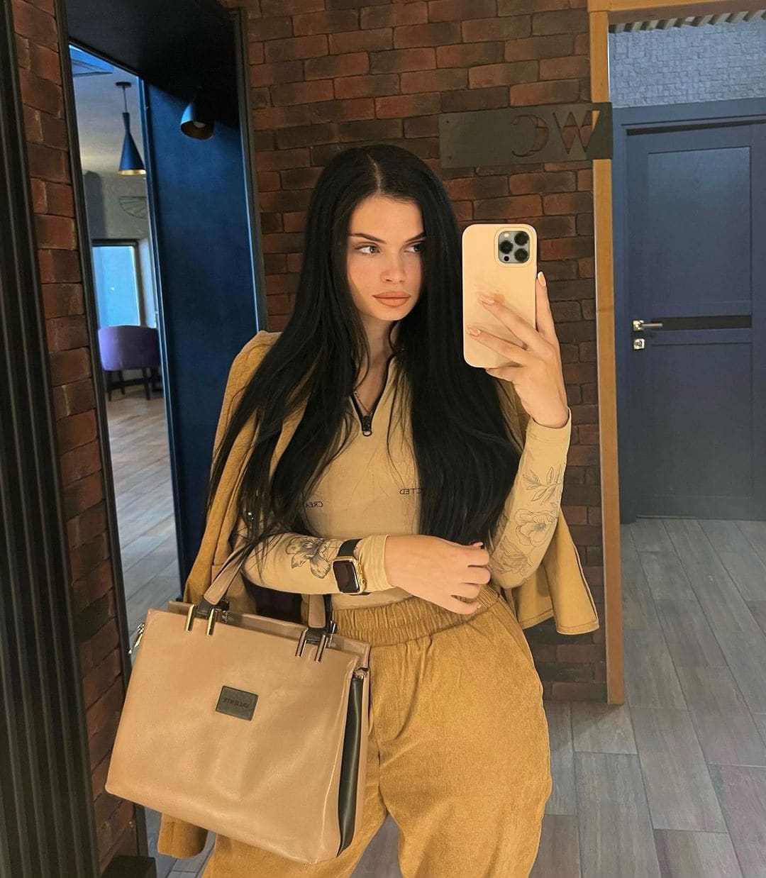 A woman takes a mirror selfie in a tan zip-neck top, trousers, and jacket, holding a large tan handbag in front of a brick wall and dark door.