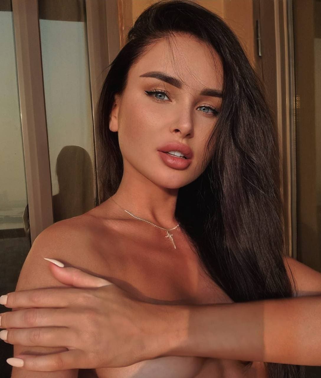Francesca with long dark hair and a cross necklace poses bare-shouldered with arms crossed near a glass door indoors.