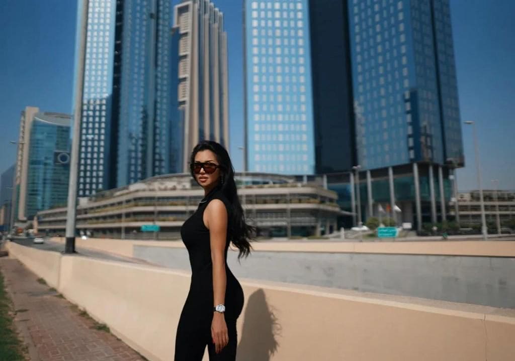 A woman in sunglasses and a black jumpsuit stands in profile on a walkway, with skyscrapers and blue sky behind her.