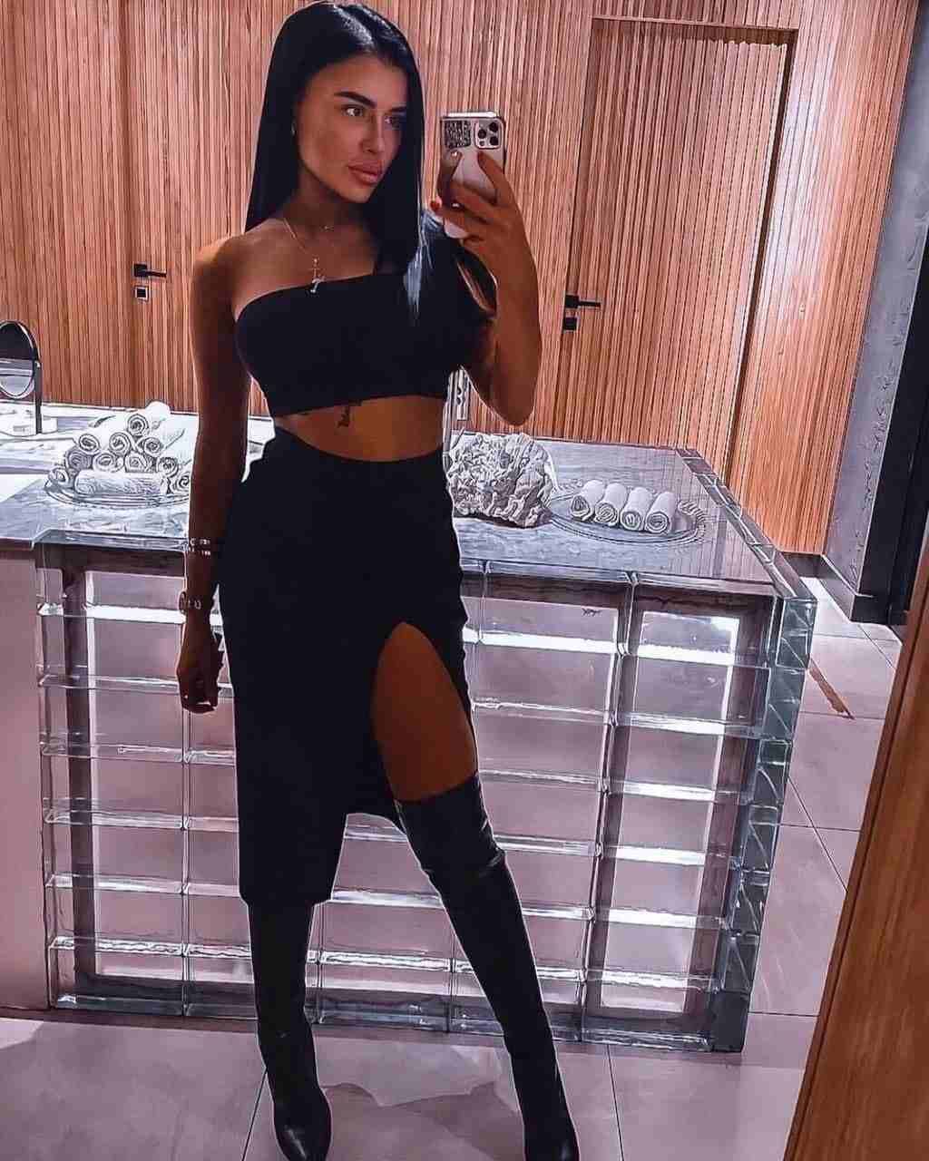 Isabel stands in a wood-paneled room wearing a black crop top, high-waisted skirt with slit, and knee-high boots, taking a mirror selfie.