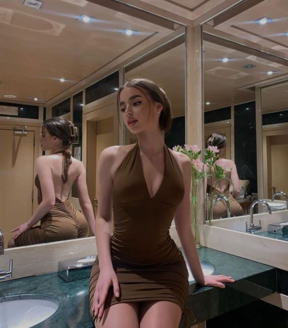 A woman in an olive-green halter mini dress sits on a marble bathroom counter, her reflection visible in the large mirrors behind her.