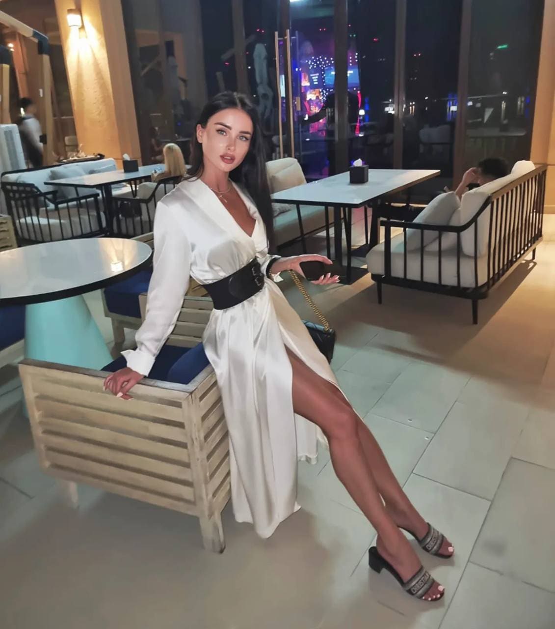 A woman in a white long-sleeved wrap dress sits on a wooden bench in a lounge, legs crossed, with a nighttime city view.