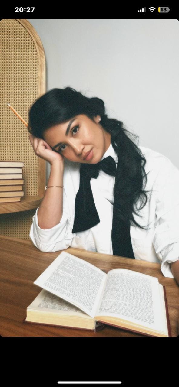 A woman with a side braid sits at a table in a white shirt and bow tie, head resting on her hand beside an open book.