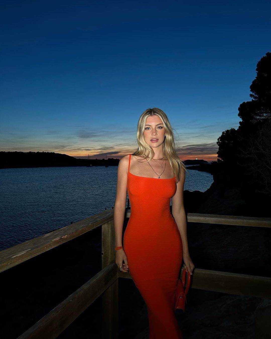 A woman stands on a wooden pier at dusk in an orange dress, holding a small bag.