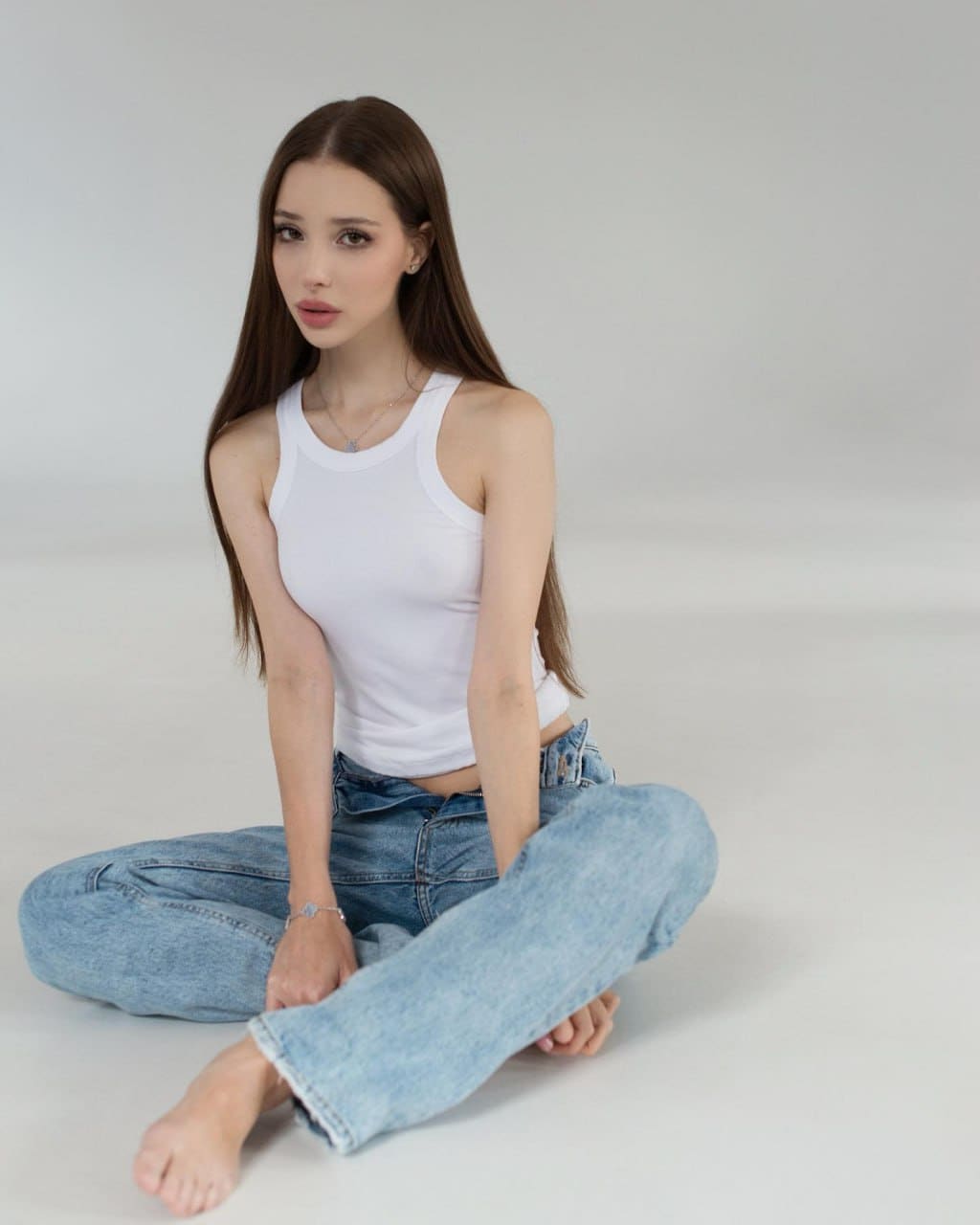 Sitting cross-legged on a light gray studio floor, a woman wears a white sleeveless tank top and light-wash blue jeans, barefoot with visible jewelry.