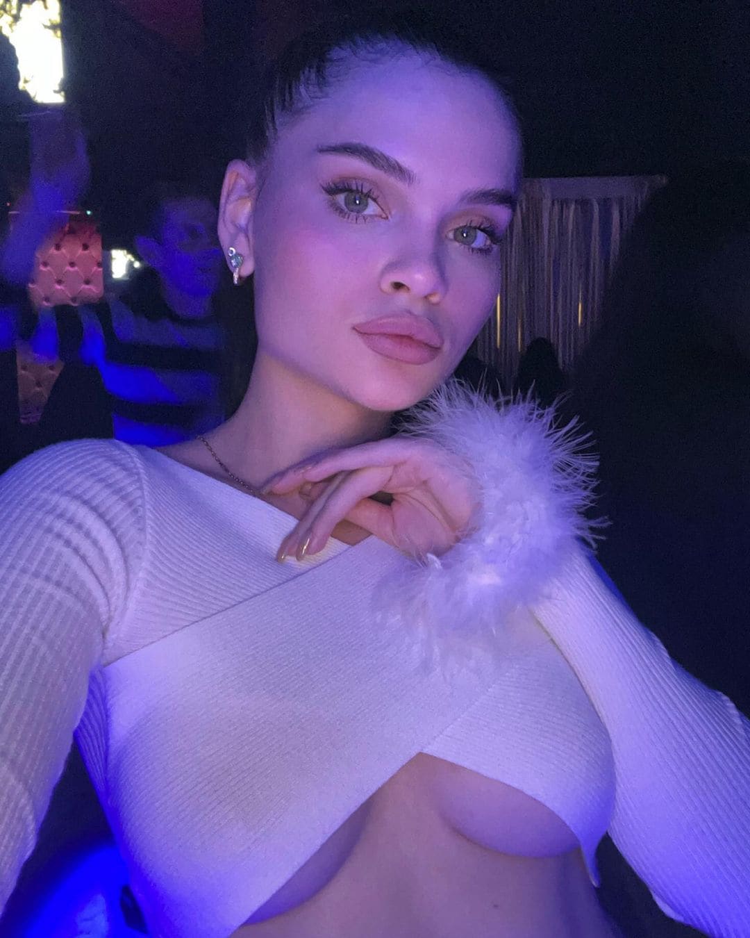 A woman poses in dim purple lighting, wearing a white ribbed long-sleeve top with a deep cutout and fluffy collar, hand near her chin.