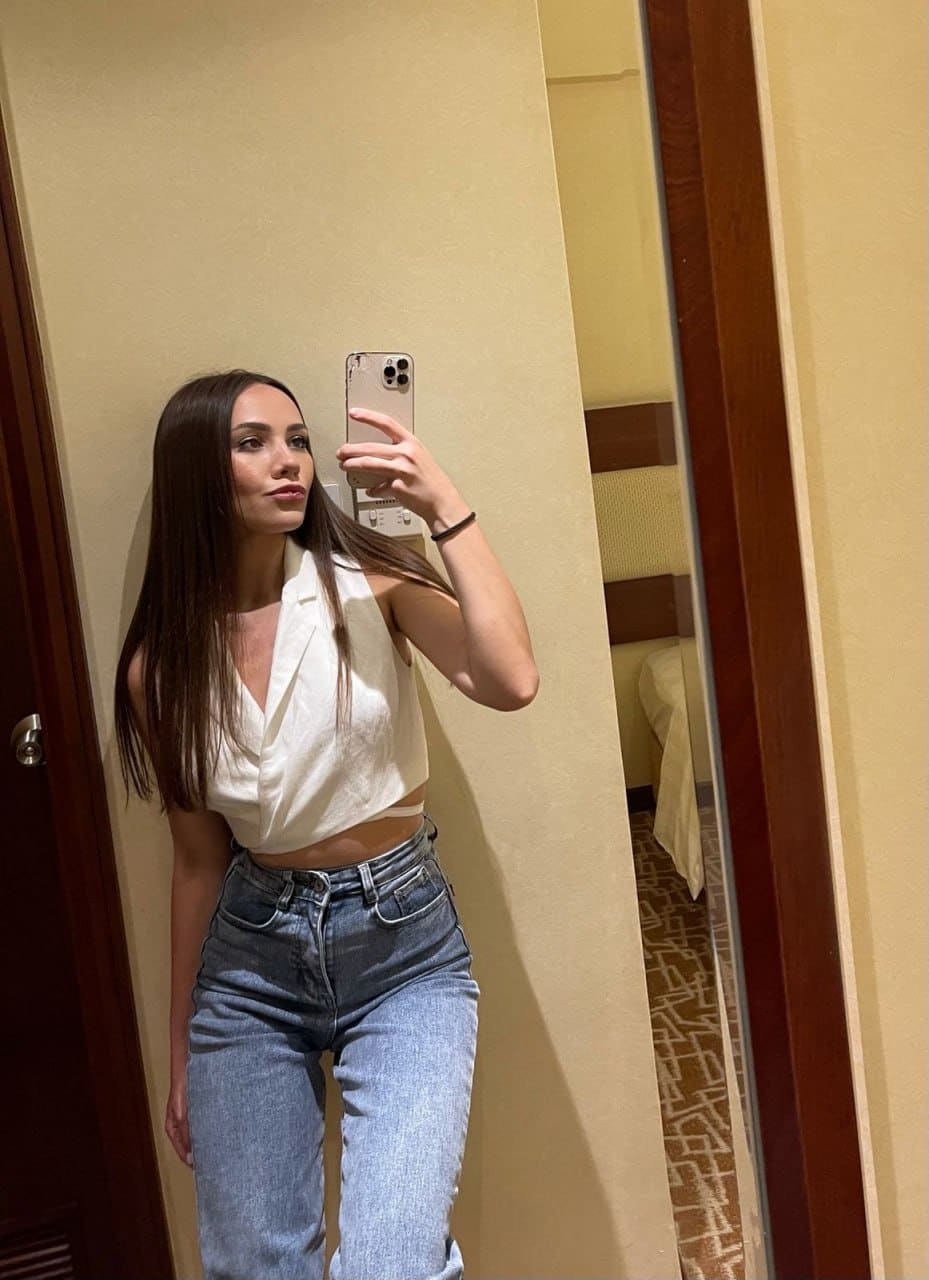 Mirror selfie in a hotel room wearing a white cropped top and light blue jeans, holding a smartphone in the right hand.