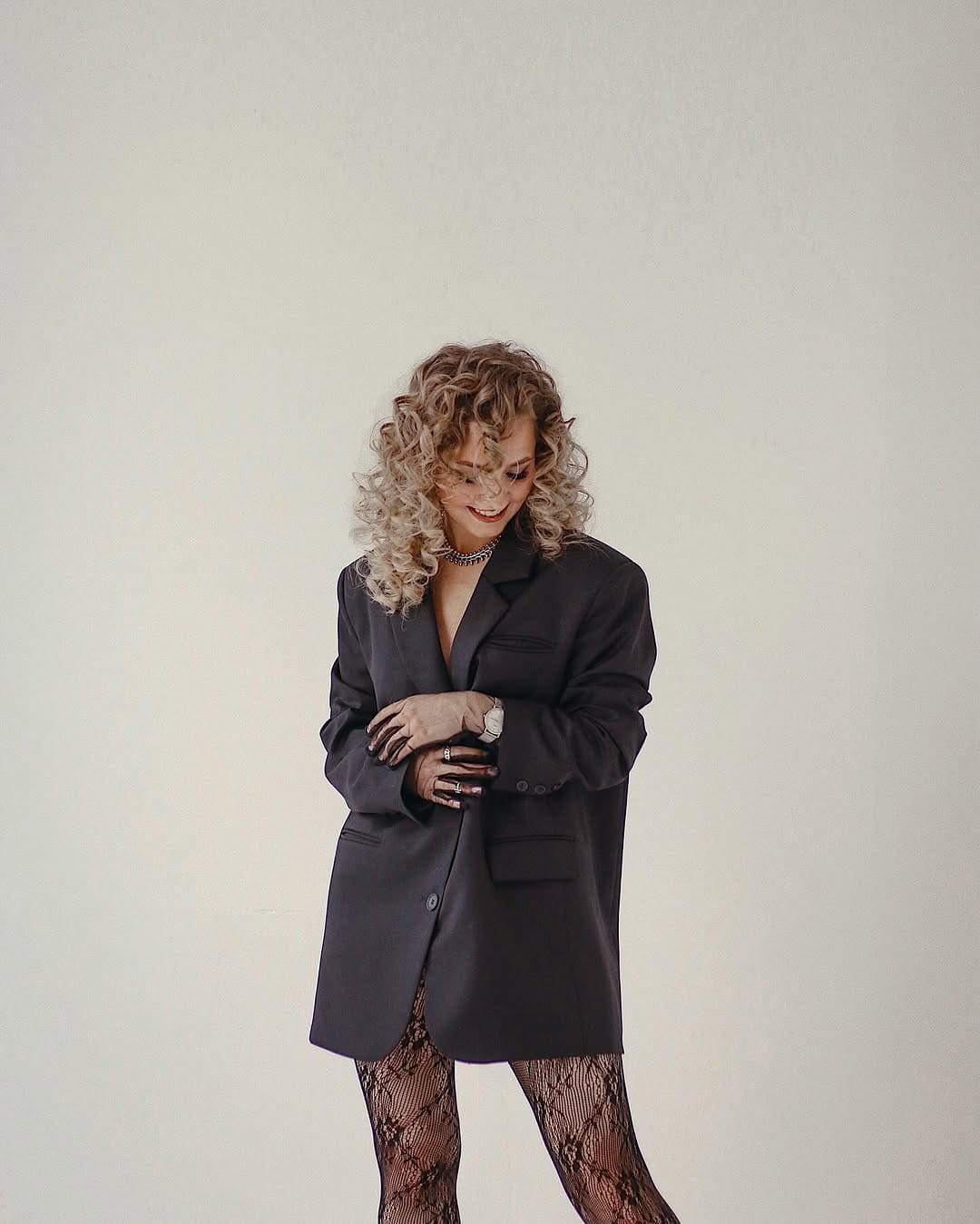 A woman stands with hands clasped wearing an oversized dark blazer, deep neckline, and patterned lace tights against a plain light wall.
