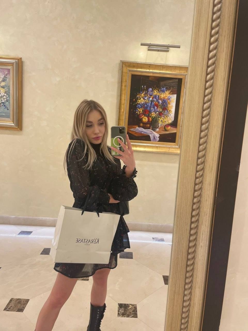 A woman stands before a mirror in a black patterned dress and lace-up boots, holding a shopping bag and smartphone in a room.