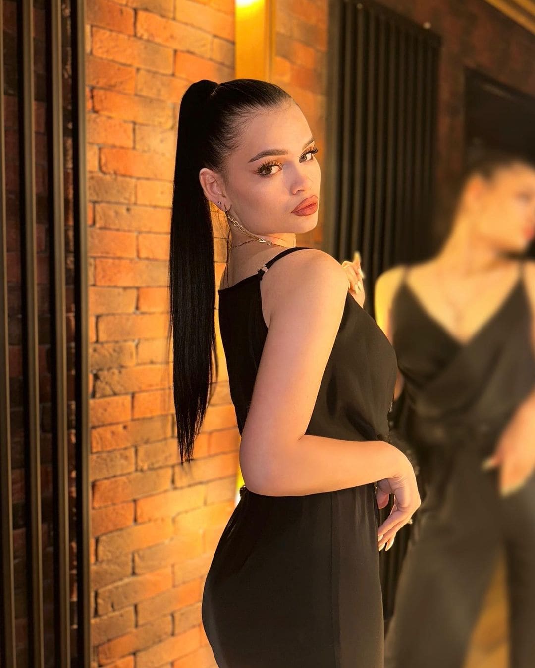 Nino stands in profile against a brick wall, wearing a sleeveless black dress with thin straps and a low back, her hand resting on her hip.