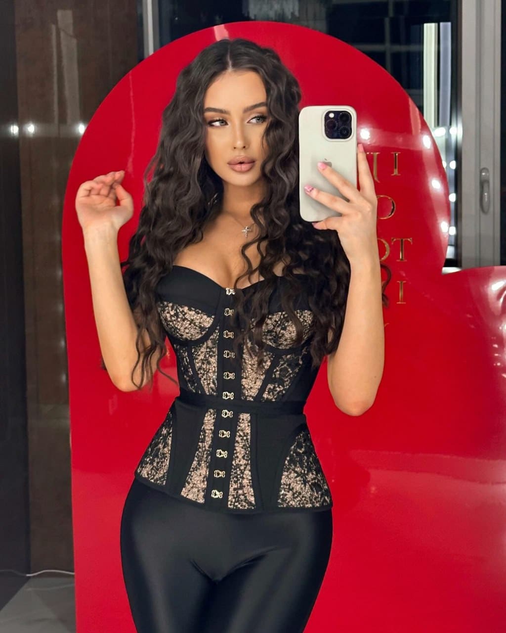 A mirror selfie shows a woman in a black lace corset and leggings standing before a large red display panel.