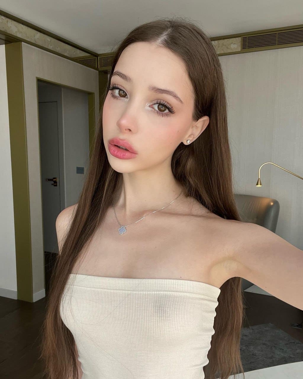 Cassandra takes a selfie in a modern interior, wearing a white strapless top and pendant necklace, bare shoulders visible near a doorway and wall lamp.