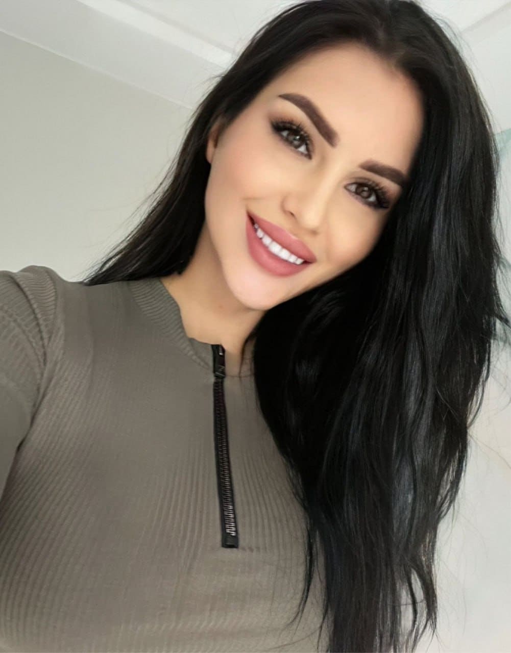 Selfie of a woman in an olive-green ribbed zip top, posed against a plain light-colored wall indoors.