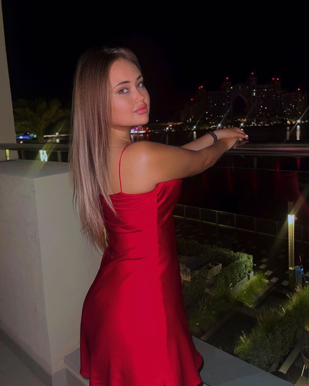 A woman stands on a balcony at night in a red satin slip dress, arms on the railing, looking back over her shoulder at a lit cityscape and water.
