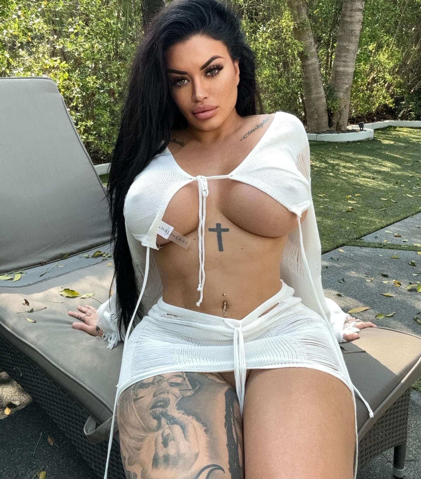 A woman sits on an outdoor lounge chair in a white open-knit top and skirt, showing thigh and chest tattoos, with green foliage behind her.